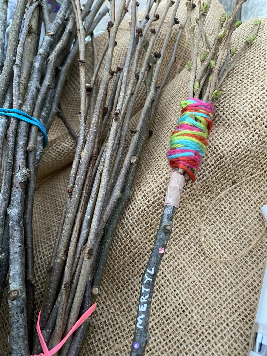 A mini broomstick made from a bundle of hazel twigs, its handle wrapped in bright coloured wool and decorated with a name, lying on hessian beside more hazel twigs