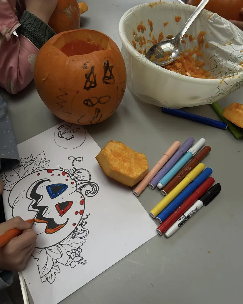 A child colours in a printed pumpkin face with felt-tip pens beside a hollowed-out pumpkin with a drawn-on face and a bowl of scooped-out pumpkin flesh